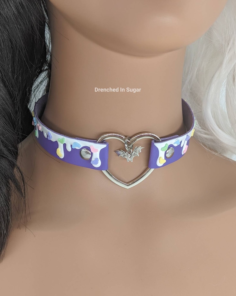 May include: Purple choker necklace with a heart-shaped charm and a bat pendant. The choker is decorated with white and pastel-colored drips and sprinkles. The text "Drenched in Sugar" is visible in the image.