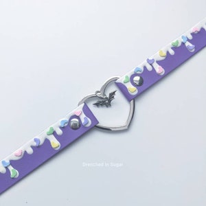 May include: Purple faux leather choker with white and pastel colored drips and a silver heart charm with a bat pendant. The text "Drenched in Sugar" is printed below the choker.