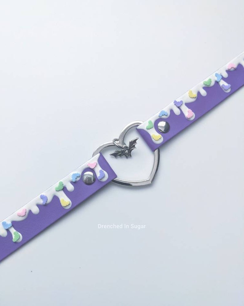 May include: Purple faux leather choker with white and pastel colored drips and a silver heart charm with a bat pendant. The text "Drenched in Sugar" is printed below the choker.