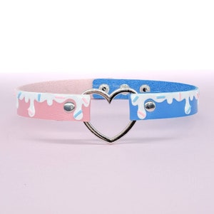 May include: A pastel-colored choker necklace with a heart-shaped center. The choker is split into pink and blue sections, each decorated with white frosting and sprinkles. The metal heart clasp adds a touch of elegance.