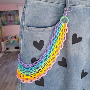 May include: A rainbow colored plastic chain hanging from a denim jean pocket. The jeans have black heart designs printed on them.