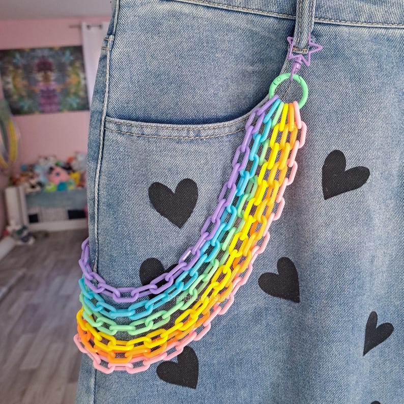 May include: A rainbow colored plastic chain hanging from a denim jean pocket. The jeans have black heart designs printed on them.