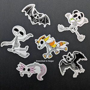 May include: Six skeleton animal stickers with black and white outlines. The stickers include a bat, a panda, a unicorn, an axolotl, and two bats. The text "Drenched In Sugar" is printed on the bottom of the image.