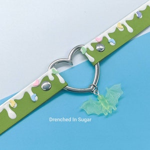 May include: Green leather choker with white icing and colorful sprinkles. A silver heart charm with a green glow-in-the-dark bat pendant hangs from the choker. The text "Drenched In Sugar" is printed below the choker.
