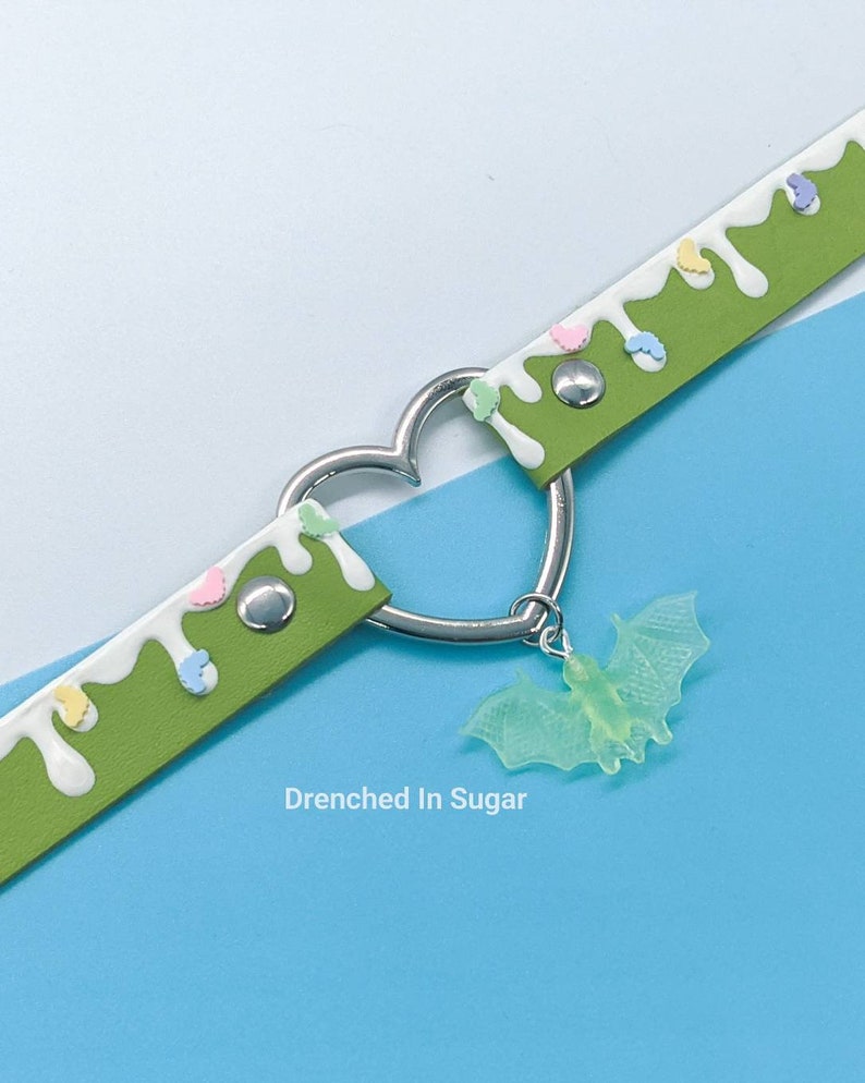 May include: Green leather choker with white icing and colorful sprinkles. A silver heart charm with a green glow-in-the-dark bat pendant hangs from the choker. The text "Drenched In Sugar" is printed below the choker.