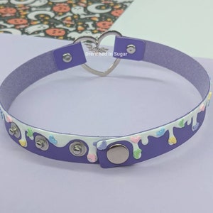 May include: Purple faux leather choker with a white drip design and colorful sprinkles. The choker has a silver heart-shaped clasp with the text "Drenched in Sugar".