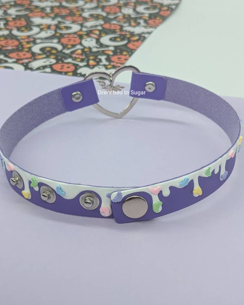 May include: Purple faux leather choker with a white drip design and colorful sprinkles. The choker has a silver heart-shaped clasp with the text "Drenched in Sugar".