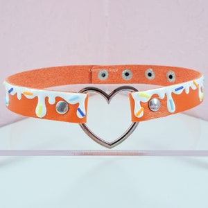 May include: Orange faux leather choker with a white icing design and colorful sprinkles. The choker has a silver heart-shaped ring.