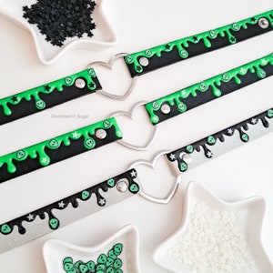 May include: Three black, silver, and green choker necklaces with heart-shaped clasps. Each necklace features a dripping green alien face design with white stars.