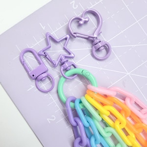 May include: A set of purple metal keychains with a star, heart, and a hook. The keychains are attached to a rainbow colored plastic chain.