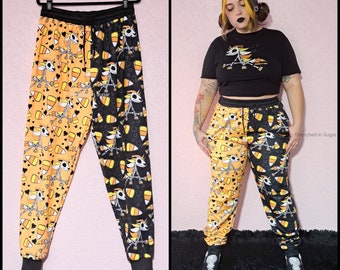 May include: Black and orange sweatpants with a skeleton candy corn design. The sweatpants are split down the middle, with one side featuring a black background and the other side featuring an orange background.