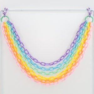 May include: A rainbow garland made of plastic chain links. The garland is hung from a white rod and is made up of seven different colors: purple, blue, green, yellow, orange, pink, and red.