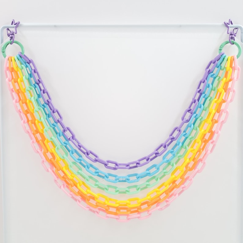 May include: A rainbow garland made of plastic chain links. The garland is hung from a white rod and is made up of seven different colors: purple, blue, green, yellow, orange, pink, and red.