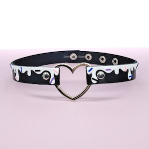 May include: Black choker necklace with a silver heart-shaped center. The black band features a white dripping design with colorful sprinkles. The choker has silver rivets and a buckle closure.