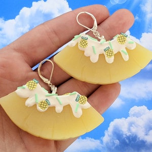 Puede incluir: Un par de pendientes amarillos en forma de piña con glaseado blanco y chispas verdes. Los pendientes están hechos de arcilla polimérica y tienen ganchos plateados.