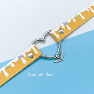 May include: A yellow leather choker with white icing and colorful sprinkles. The choker has a heart-shaped clasp and a silver bat charm.