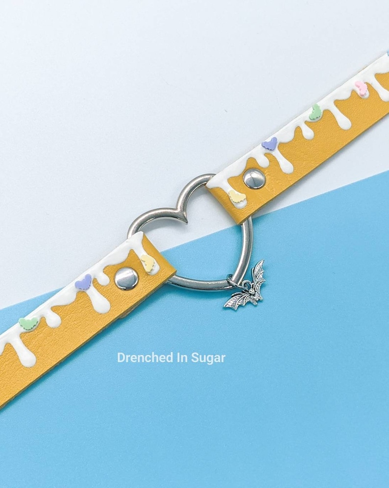 May include: A yellow leather choker with white icing and colorful sprinkles. The choker has a heart-shaped clasp and a silver bat charm.
