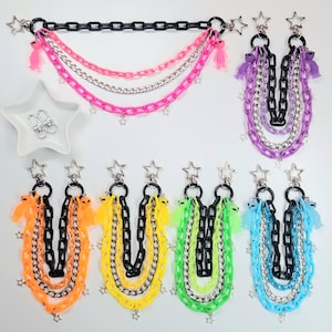 May include: A collection of colorful chain accessories, including keychains and a chain belt. The keychains feature black, silver, and colored chains with star charms and alien-shaped charms. The chain belt has pink, silver, and black chains with star charms and alien charms.