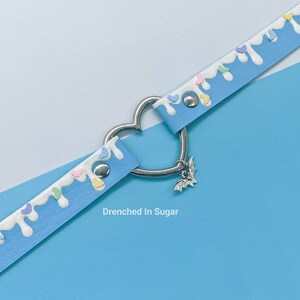 May include: A light blue choker necklace with white icing and colorful sprinkles. The necklace has a heart-shaped metal clasp and a silver bat charm. "Drenched In Sugar" is written below the necklace.