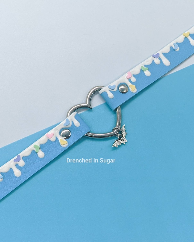 May include: A light blue choker necklace with white icing and colorful sprinkles. The necklace has a heart-shaped metal clasp and a silver bat charm. "Drenched In Sugar" is written below the necklace.