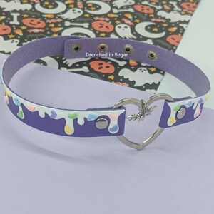 May include: Purple faux leather choker with a white drip design and colorful sprinkles. The choker has a silver heart-shaped clasp with a bat charm.