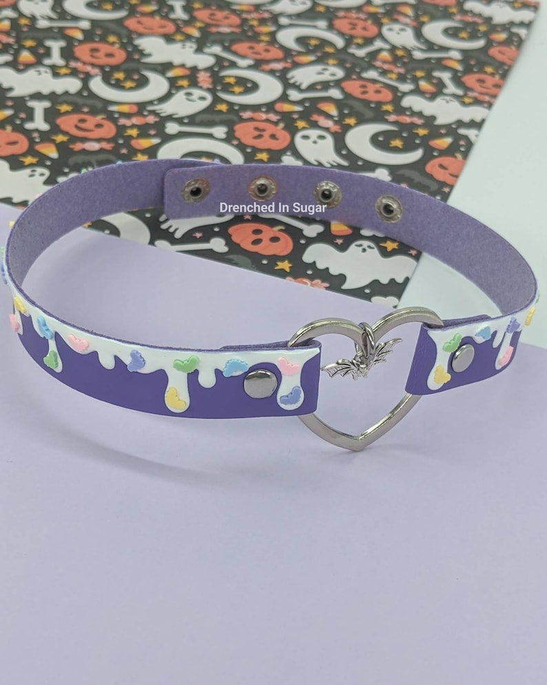 May include: Purple faux leather choker with a white drip design and colorful sprinkles. The choker has a silver heart-shaped clasp with a bat charm.