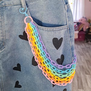 May include: A rainbow colored plastic chain with a silver clip attached to a pair of blue jeans. The chain is hanging from the front pocket of the jeans.