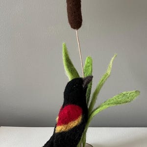 May include: A handcrafted felt bird with black feathers, a red and yellow wing patch, and a dark beak, perched on green felt leaves and a cattail. The bird is mounted on a small wooden base, creating a nature-inspired decorative piece.