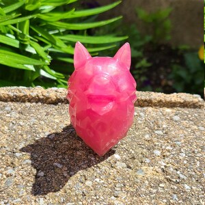 May include: A pink resin wolf head sculpture with a geometric design. The sculpture is sitting on a rough, textured surface.
