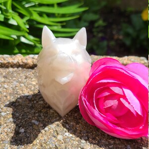 May include: A white ceramic figurine of a cat with a pink flower next to it. The cat figurine has a shiny, pearlescent finish.