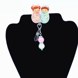 May include: A badge reel with two cartoon babies in pastel-coloured bunny-eared blankets. The reel has a clear plastic clip and a beaded charm with pink and pastel-coloured beads. The babies have pacifiers and are decorated with Easter-themed images.