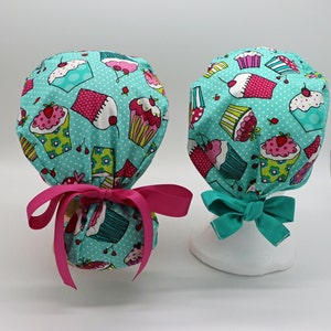 May include: Two turquoise blue surgical caps with a pink and white cupcake pattern. The caps have pink and turquoise blue ties.