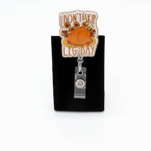 May include: A badge reel featuring a decorative topper with a roasted turkey illustration, sunflowers, and the text I DON'T SKIP LEG DAY. The topper has a glittery border and is attached to a clear plastic strap with a metal snap button closure.