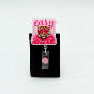 May include: A badge reel featuring a rectangular acrylic topper with a tiger wearing pink heart-shaped sunglasses and a pink feather boa. The word easy is printed in pink script above the tiger. A clear plastic strap with a pink glitter button.