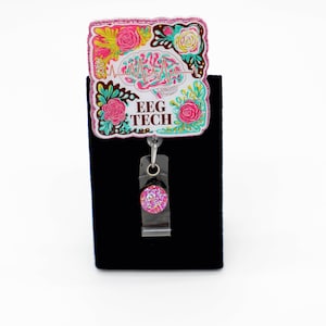 May include: A badge reel featuring a rectangular topper with a colorful illustration of a brain, a heartbeat line, and floral accents in pink, teal, and yellow. The text EEG TECH is printed in brown. A small, circular, pink glittery accent is attached.