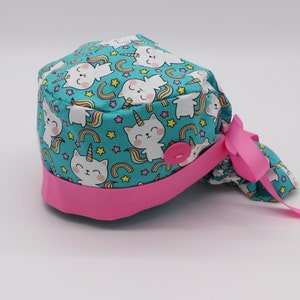 May include: A turquoise scrub hat with a pink trim and a button closure. The hat features a repeating pattern of white cats with unicorn horns and rainbows.