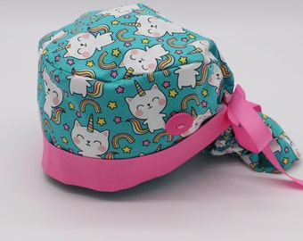May include: A turquoise scrub hat with a pink trim and a button closure. The hat features a repeating pattern of white cats with unicorn horns and rainbows.