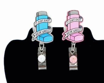Op de afbeelding: Twee badgehouders met acryl inhalatorontwerpen en glitterranden. De ene inhalator is blauw, de andere roze. Beide tonen een wit lint met de zwarte tekst IT AINT EASY BEIN' WHEEZY. Elke houder heeft een doorzichtig plastic riempje en een kleine ronde glitteraccent.