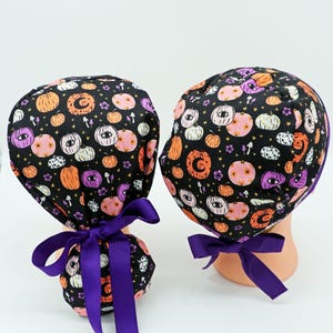 Może przedstawiać: Dwa czarne czepki chirurgiczne z wzorem dyni na Halloween w kolorze pomarańczowym, różowym i białym. Każdy czepek ma fioletową wstążkę zawiązaną z tyłu w kokardę. Czepki są prezentowane na białym tle.