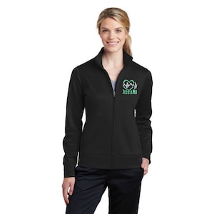 Puede incluir: Chaqueta negra con cremallera y cuello alto. La chaqueta presenta un gráfico en forma de corazón verde y blanco con el texto "JESSARI" en el pecho izquierdo. La chaqueta se lleva sobre una camiseta gris y pantalones negros.
