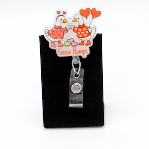 May include: A badge reel with a cartoon design featuring two geese holding hands, wearing sweaters and heart-shaped balloons. The text "Goose-Bumps" is below the geese. The reel has a clear plastic strap and a metal snap.