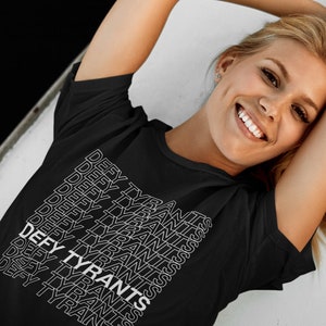 May include: A woman wearing a black t-shirt with the text "DEFY TYRANTS" repeated in white lettering.