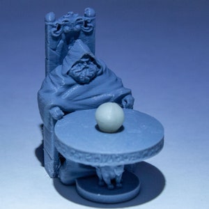 Könnte beinhalten: Eine blaue 3D-gedruckte Miniatur einer robenbekleideten Figur, die auf einem Thron sitzt, mit einer Kristallkugel auf einem Tisch vor ihr.