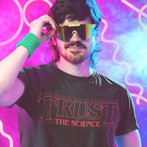 Puede incluir: Un hombre con una camiseta negra con el texto "TRUST THE SCIENCE" en rojo. Lleva pantalones verde neón y gafas de sol futuristas. El fondo es un degradado morado y azul con luces de neón.