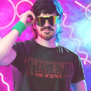 Puede incluir: Una persona con una camiseta negra con el texto "TRUST THE SCIENCE" en rojo. Lleva pantalones verde neón y gafas de sol. El fondo es un degradado púrpura y azul con luces de neón.
