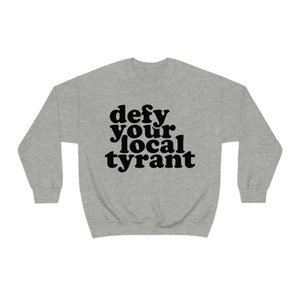May include: A heather gray sweatshirt with the phrase "defy your local tyrant" printed in bold, black letters. The sweatshirt has a crew neck and long sleeves, suitable for casual wear.