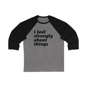 Puede incluir: Una camiseta de béisbol gris y negra con la frase "i feel strongly about things" impresa en negro. La camiseta tiene mangas negras y un cuerpo gris, ofreciendo un estilo casual y cómodo.