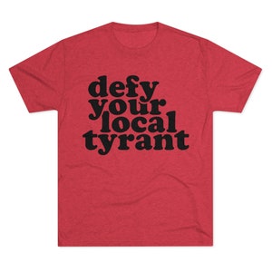 May include: Red t-shirt with the phrase "defy your local tyrant" in bold, black letters. The shirt has a crew neck and short sleeves. The fabric has a heathered texture.