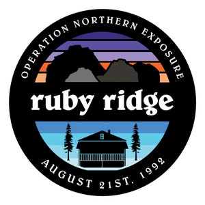 May include: A black and white circular sticker with a sunset scene, mountains, a cabin, and trees. The text "Operation Northern Exposure ruby ridge August 21st, 1992" is printed on the sticker.