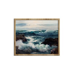 May include: A framed wall art print depicting a turbulent ocean scene with deep blue and teal waves crashing against dark brown rocky outcrops under a soft, overcast sky. The artwork is enclosed in a light-toned wooden frame with a visible grain.